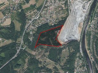 Ortofotomapa zobrazující zalesněnou oblast označenou červenou polygonovou hranicí, sousedící s velkým lomem/těžební oblastí na východě a zástavbou na severu.