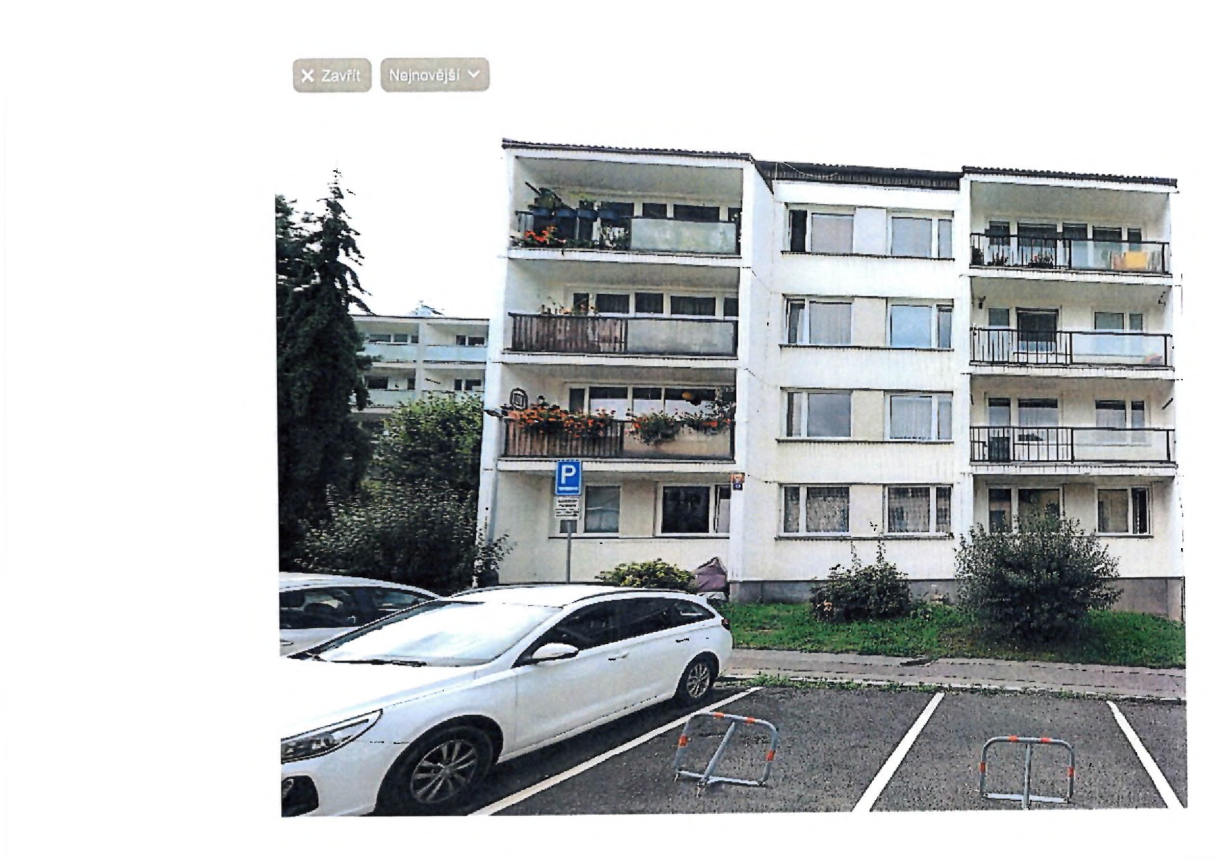 Fotografie bytového domu s parkovištěm. Na budově je umístěna modrá dopravní značka 'P' (parkoviště) s dodatkovou tabulkou, jejíž text je nečitelný.