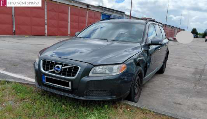 Fotografie poškozeného tmavě šedého osobního automobilu značky Volvo (kombi) zaparkovaného na zpevněné ploše před řadou garáží. V levém horním rohu je logo 'F. FINANČNÍ SPRÁVA'.