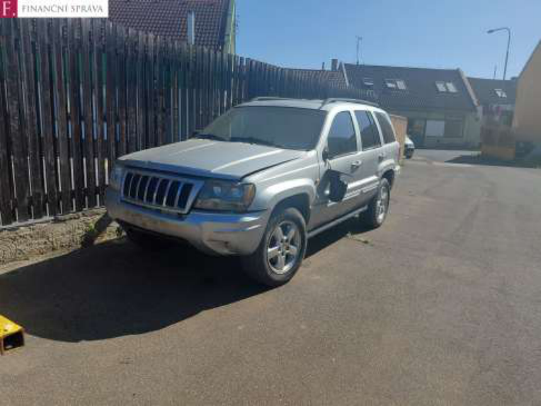 Fotografie stříbrného osobního automobilu značky Jeep (model Grand Cherokee) zaparkovaného u dřevěného plotu. V levém horním rohu je logo 'F. FINANČNÍ SPRÁVA'.