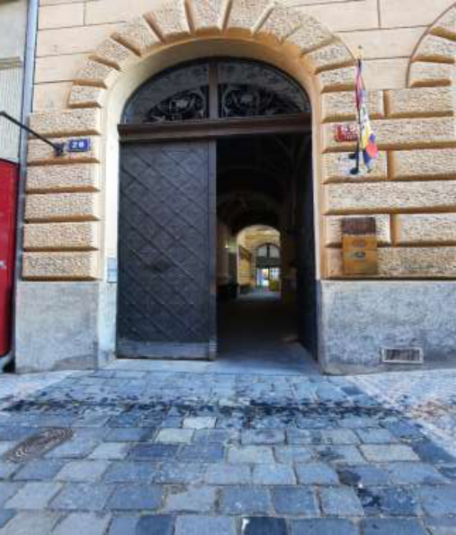 Fotografie budovy s vchodem do průjezdu. Na budově je umístěna modrá orientační tabulka s číslem popisným '26'. Dále je na budově umístěna vlajka a informační tabule (text na tabuli není čitelný).
