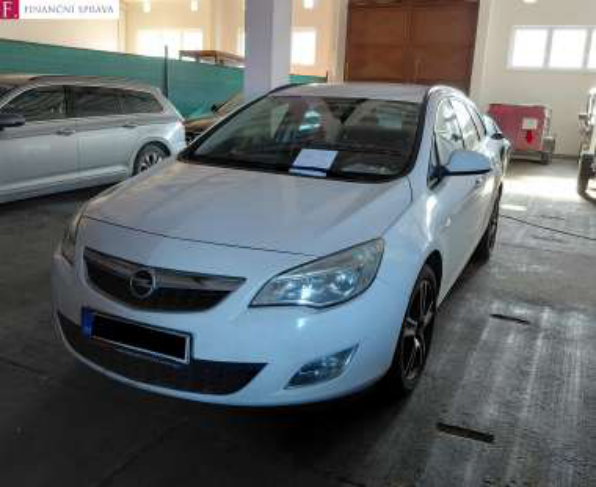 Fotografie bílého osobního automobilu značky Opel (model Astra J) v interiéru haly. V levém horním rohu je logo 'FINANČNÍ SPRÁVA'. Na čelním skle vozu je umístěn papírový štítek.