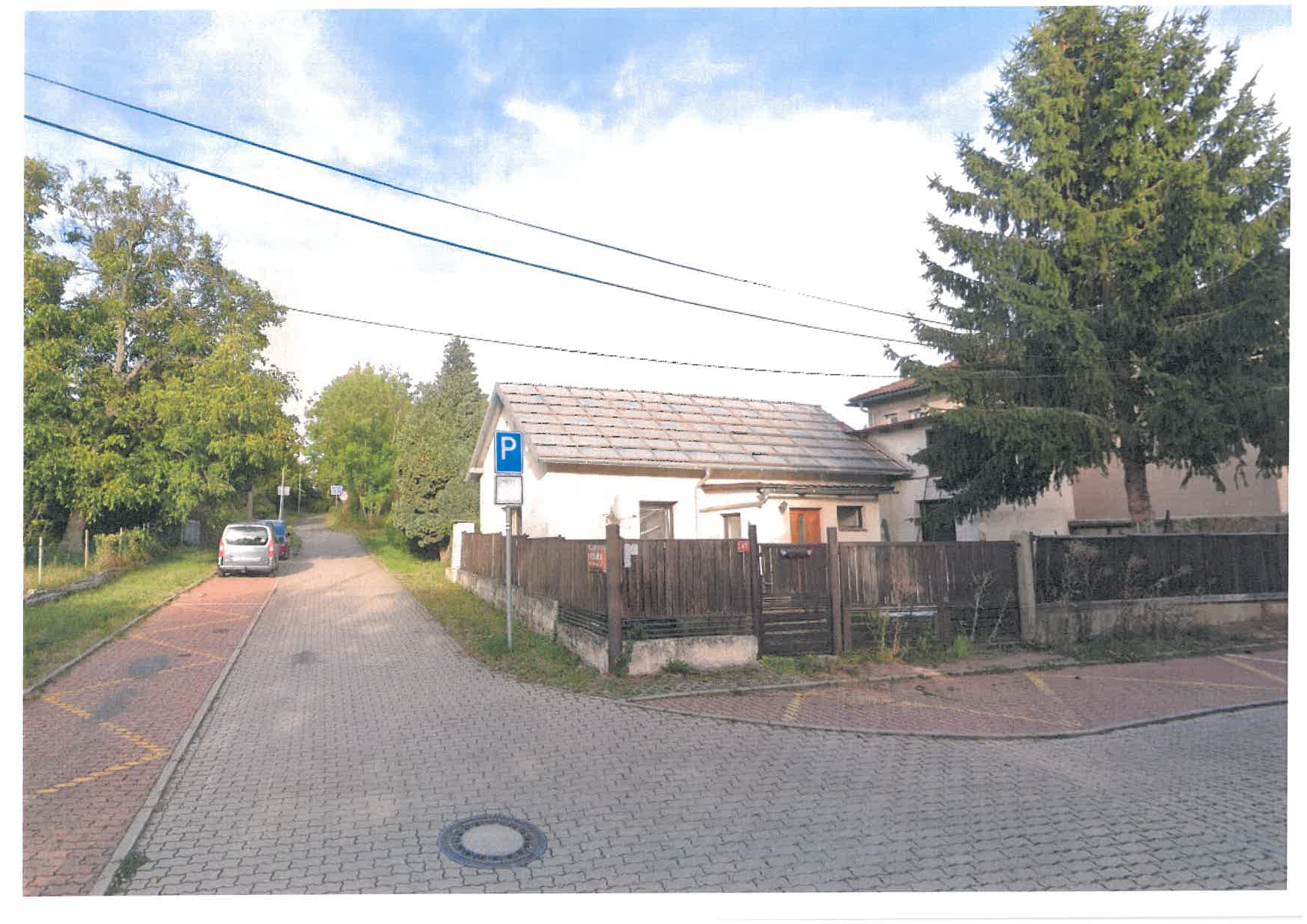 Fotografie budovy a přilehlé ulice. Na budově je umístěna modrá dopravní značka 'P' (parkoviště) s doplňkovou tabulkou, na které je čitelné číslo popisné '15'.