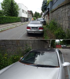 Fotografie ulice s parkujícími auty (stříbrný Mercedes-Benz a černé vozidlo) podél kamenné zdi a rodinných domů.
