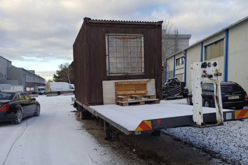 Fotografie zachycující dřevěnou buňku naloženou na přívěsu. Na černém osobním automobilu vpravo je čitelná registrační značka (SPZ): 9P0 0647.