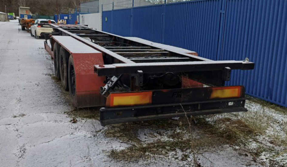 Fotografie nákladního návěsu (kontejnerového podvozku) značky Fliegl odstaveného na zpevněné ploše. Na zadním nárazníku je částečně čitelný nápis 'Fliegl'.