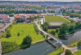 Vizualizace architektonického projektu v městské zástavbě. Obrázek zachycuje návrh nové budovy s plochou zelenou střechou situovanou vedle historického průmysl…