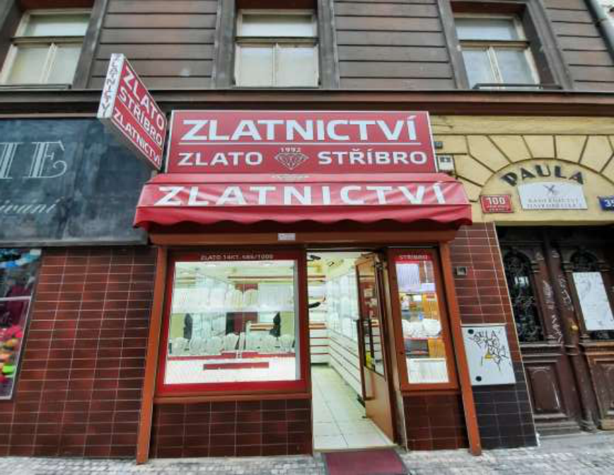 Fotografie průčelí obchodu se šperky. Na vývěsním štítu a markýze je nápis 'ZLATNICTVÍ', 'ZLATO', 'STŘÍBRO' a rok '1992'. V okně je nápis 'ZLATO 14KT 585/1000'. Vedle vchodu je cedule s nápisem 'PAULA' a číslo popisné '100'.