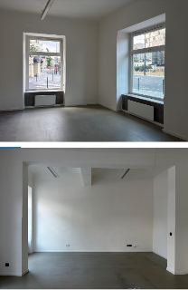 Fotografie interiéru prázdných komerčních prostor s bílými stěnami, šedou podlahou a velkými okny s výhledem na ulici. Na stěnách jsou patrné elektrické zásuvk…