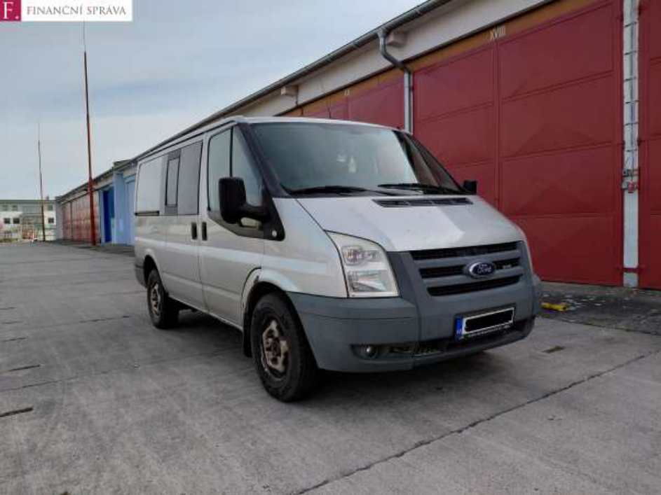 Fotografie stříbrné dodávky značky Ford (model Transit) zaparkované před budovou s červenými vraty. V levém horním rohu je logo 'F. FINANČNÍ SPRÁVA'.