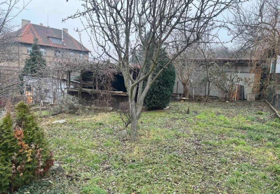 Fotografie pozemku se zahradní stavbou a vzrostlou zelení.