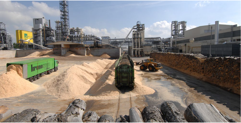 Fotografie průmyslového areálu (pravděpodobně dřevozpracující závod), na zeleném nákladním voze je čitelný nápis 'kronospan'.