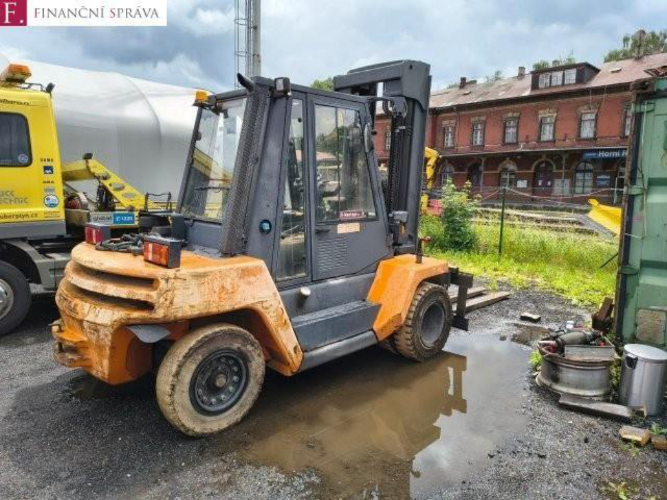 Fotografie vysokozdvižného vozíku oranžovo-šedé barvy stojícího na venkovní ploše. V levém horním rohu je logo 'F. FINANČNÍ SPRÁVA'. Na pozadí je vidět cihlová budova s nápisem 'Horni R' a žlutý nákladní vůz s částečně čitelným textem '...uberplyn.cz'.