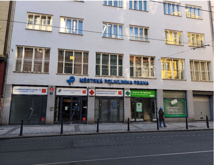 Fotografie budovy Městské polikliniky Praha. Na budově jsou nápisy: 'MĚSTSKÁ POLIKLINIKA PRAHA', 'ODBĚROVÉ MÍSTO COVID-19', 'PŘÍSPĚVKOVÁ ORGANIZACE HLAVNÍHO MĚSTA PRAHY' a 'Lékárna Ve Spálené'. Dále je zde vidět označení vchodu a provozní informace na dveřích.