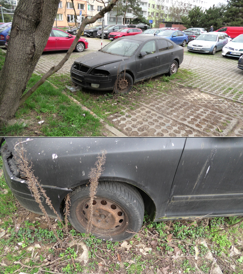 Fotografie opuštěného, nepojízdného černého osobního automobilu značky Škoda (pravděpodobně model Octavia) zaparkovaného na parkovišti s betonovými zatravňovacími dlaždicemi. Vozidlo je v zanedbaném stavu, chybí mu přední maska a světlomety, má zrezivělé disky kol a je částečně zarostlé vegetací.