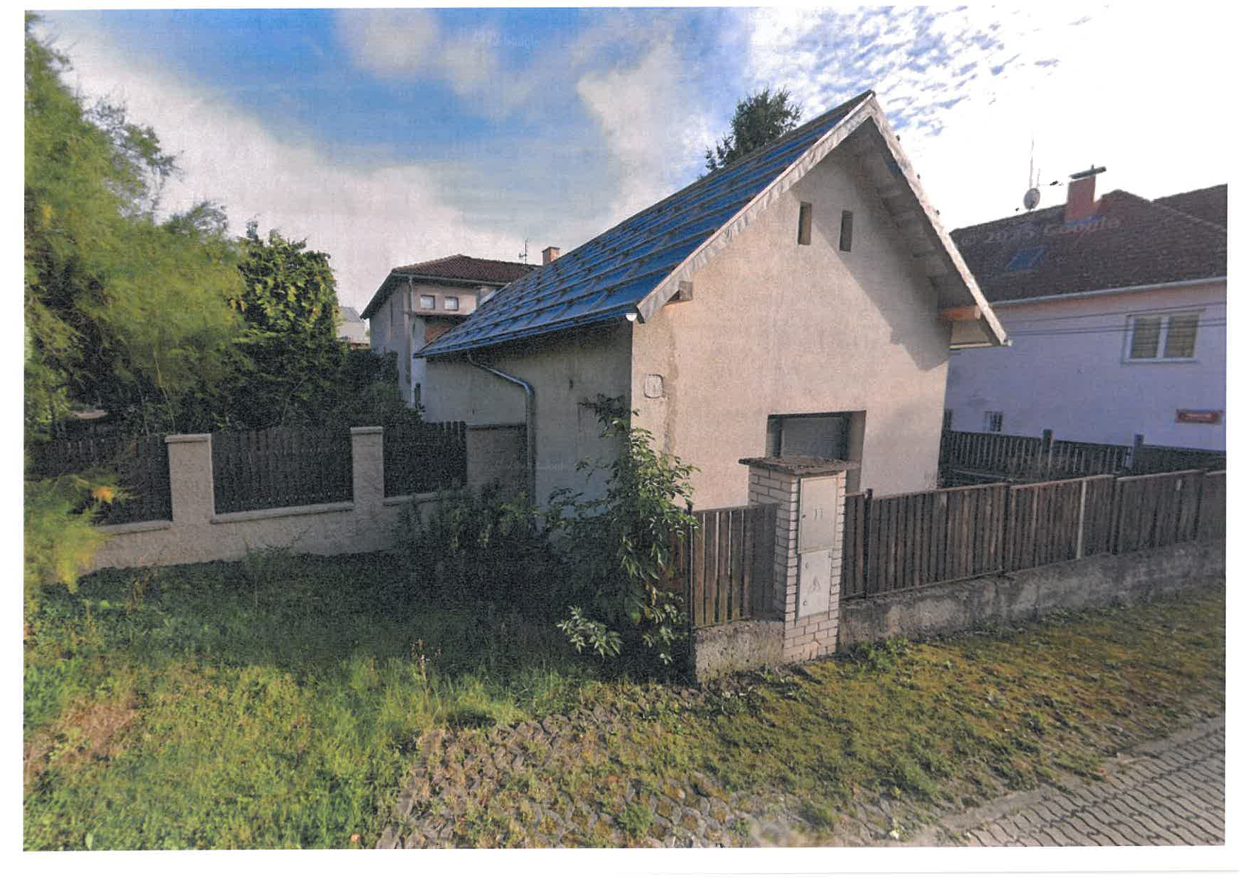 Fotografie exteriéru rodinného domu se sedlovou střechou, oplocením a elektrickým rozvaděčem na sloupku u vstupu na pozemek.