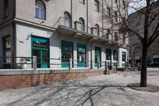 Fotografie budovy s obchody (vlevo s nápisem 'BACKYARD' nad vstupem) a dlážděným náměstím se stromem v popředí.