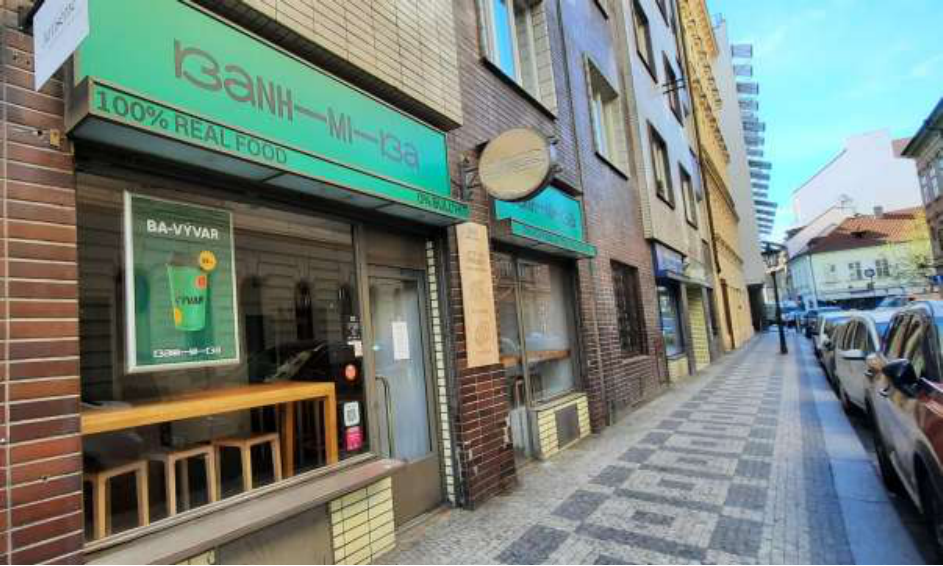 Fotografie provozovny 'BANH-MI-BA' s nápisem '100% REAL FOOD'. V okně je umístěn plakát na produkt 'BA-VYVAR'.