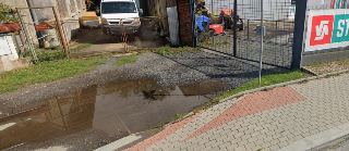 Fotografie pozemku a vjezdu s velkou kaluží vody na příjezdové cestě před branou do areálu.
