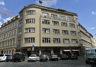 Fotografie budovy na rohu ulice s obchody v přízemí, parkujícími auty a dopravními značkami.