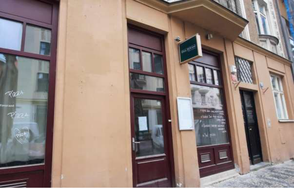 Fotografie průčelí budovy s restaurací. Na oknech je nápis 'PIZZA' a na vývěsním štítu nad vchodem je nápis 'PIZZA DI VINO'. Na pravém okně je ručně psaný text s nabídkou (menu), který je však z větší části nečitelný.